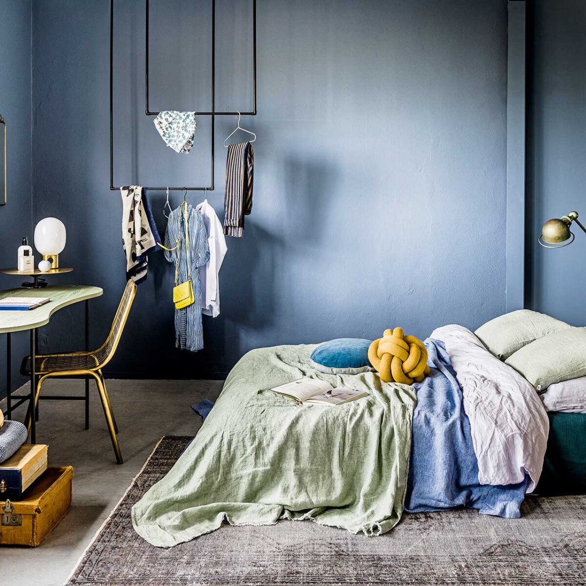 Juniper Ash 115, absolute matt emulsion (Little Greene). Twee matrassen (IKEA). Linnen donkergroen dekbedovertrek als onderlaken, denimblauw dekbedovertrek (Sissy-Boy). Stonewashed dekbedovertrek (Puur Linnen). Jadegroene plaid van By Mölle, velours rond kussen met gouden piping van Bloomingville, wit-blauwe tie-dye-vaas van Lifestyle, tafellamp op goudkeurige voet van Menu, messing draadstoel (via vtwonen). Kelim (Rozenkelim). Okergeel kussen Knot van Design House Stockholm (misterdesign.nl). Houten bankje achter het bed, spiegel koperen dienblad, borden (De Weldaad). Geel schaaltje, kaars (Six and Sons). Boeken (kringloop). Vintage Jieldé-lamp privébezit. Behang Byron van Cole &amp; Son (De Mooiste Muren). DIY-kledingrek van waterleidingbuizen (Gamma). Kledinghangers per 5 (Hay). Bureau met goudkleurige verhoging (Mobilia). Koffers (Burbri Antiek). Plaid (By Mölle). Schoenen (Love Stories). Sjaal (Six and Sons). Witte blouse, broek met streep (Sukha Amsterdam). Kan (kringloop). Beautyproducten privébezit. vtwonen 09-2017 | Fotografie Sjoerd Eickmans | Styling Leonie Mooren, Liza Wassenaar