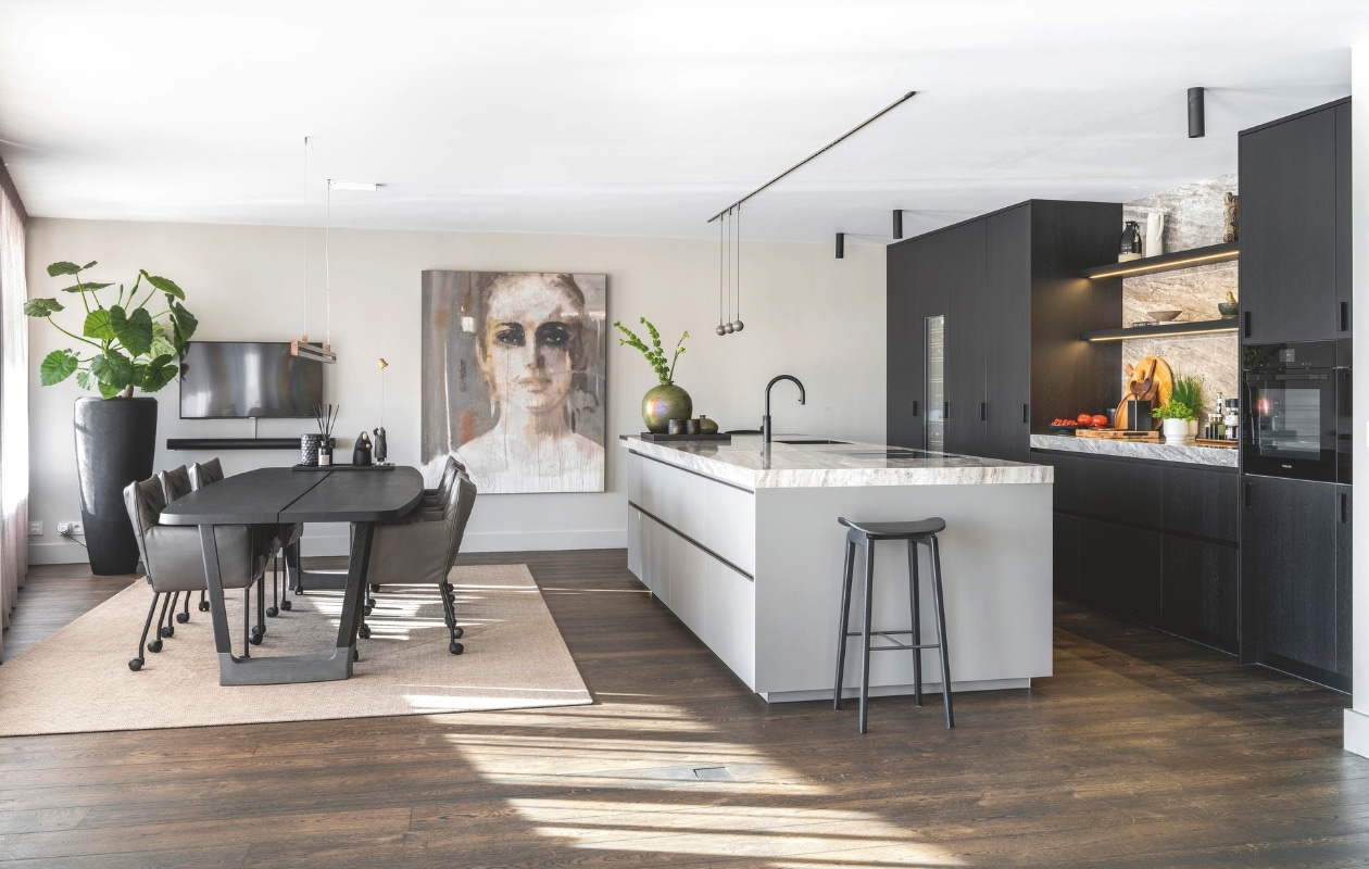 De eettafel van Arco is bij Meijs Wonen aangeschaft. De stoelen zijn van Label, het karpet van Limited Edition, alles door Raw Interiors geleverd. De grote pot met plant komt van Raw Interiors. Het schilderij is van de Bossche kunstenaar Jos van Uitregt. De keuken is ontworpen en gerealiseerd door Van Boven. Het werkblad is van keramiek. De barkruk is Deens design van Norr11. De lampen boven het werkblad komen van De Bonth Licht &amp; Advies. stijlvol wonen 10-2024 | fotografie Denise Zwijnen