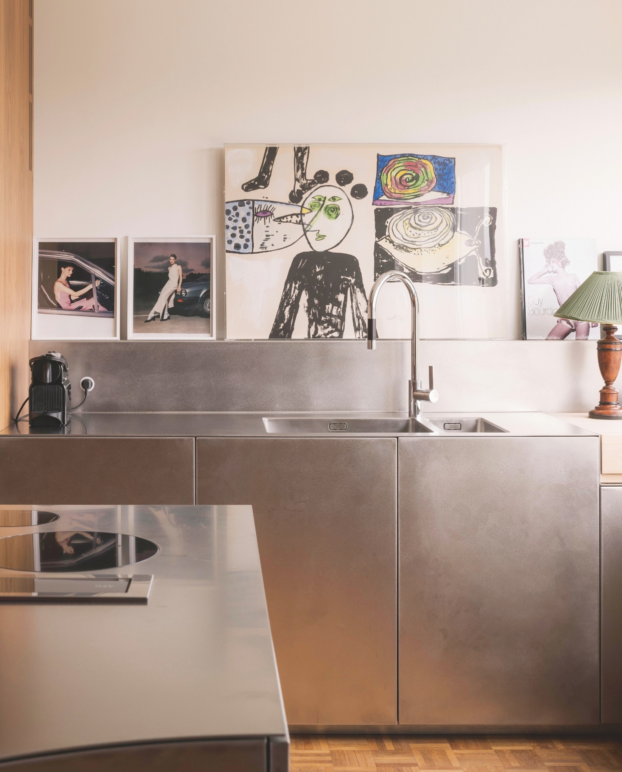 De koele look van de keuken wordt gecompenseerd door het warme hout en de kleurrijke kunstwerken. Tegen de muur: twee foto’s van Studio Soir-campagnes naast een schilderij van Corneille. stijlvol wonen 07-2024 | fotografie Katoo Peeters