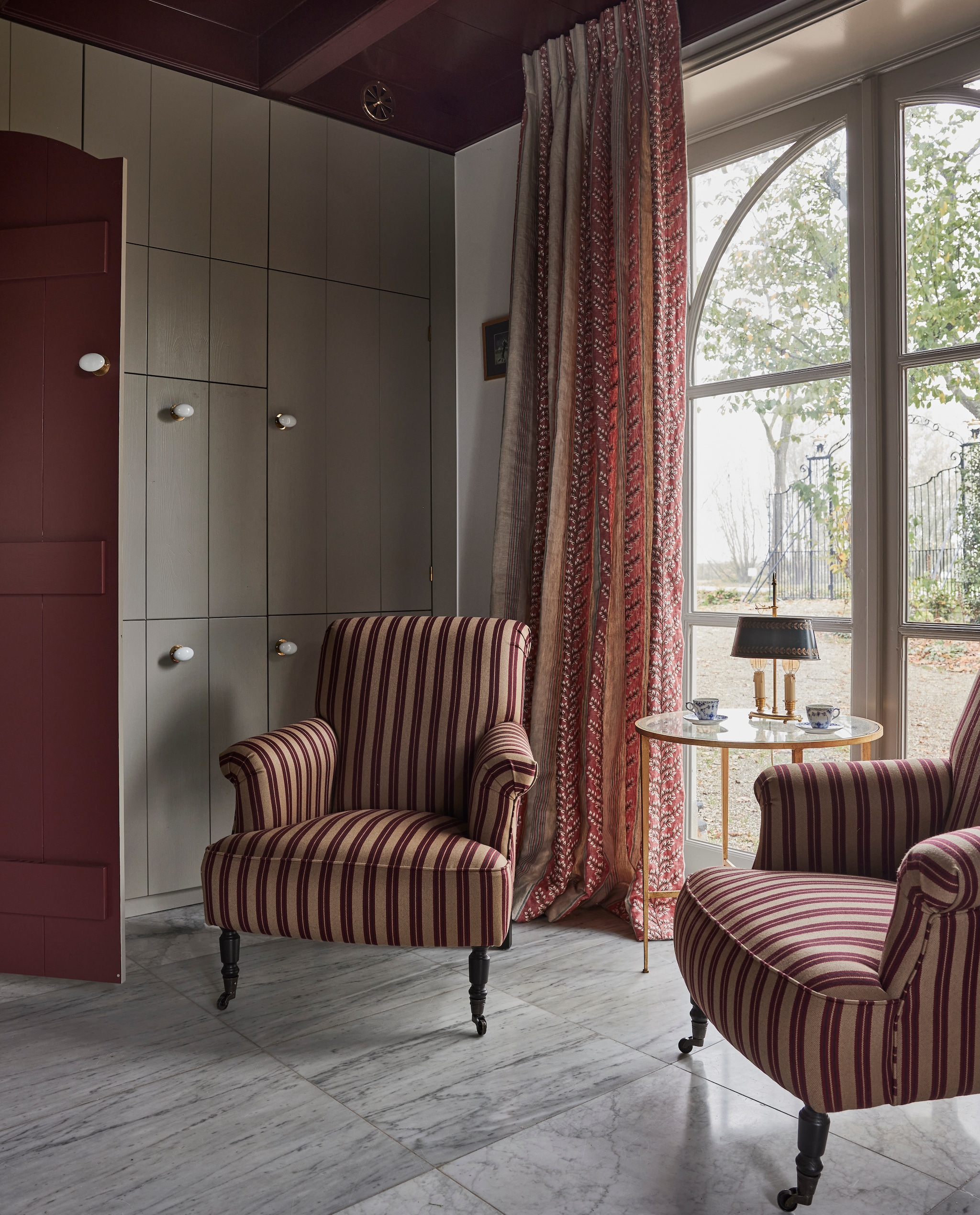 Fauteuil Bonifacio in stof Rayure Gladstone Nobilis, via Wilhelmine van Aerssen Agenturen. Bijzettafel Vaughan Designs. Antieke Bouillot tafellamp. Kastenwand op maat gemaakt door Hans Tinga. Kleur houtwerk Persiaans rood vanVettewinkel. vtwonen landelijk 01-2025 | styling Marianne Luning | fotografie Alexander van Berg | tekst Merel van der Lande