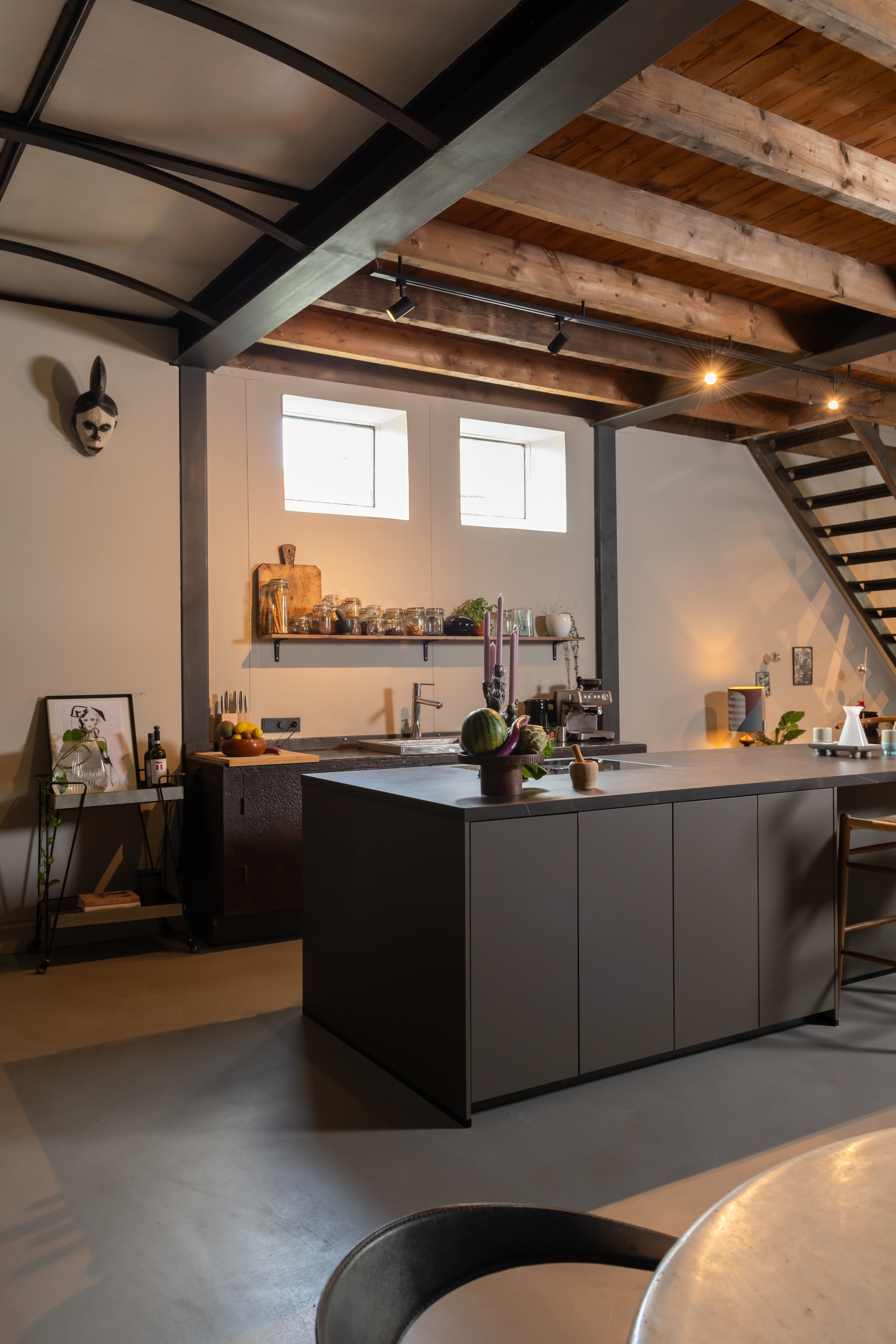 Betonnen vloer origineel, kookeiland Clear Kitchen, houten plank Praxis, broodplank van een markt in Frankrijk, potten met zaden Ikea. Marokkaanse glaasjes en fruitschaal Soft Interieur. Kandelaar zelfgemaakt door Margot, messenblok Kuhn Rikon. Bruine schaal en witte vaas vintage. Masker aan de muur meegenomen uit Zuid-Afrika, barkruk Sklum. Eetkamerstoel en ronde tafel Marktplaats. vtwonen 02-2025 | Fotografie Caroline Martinot