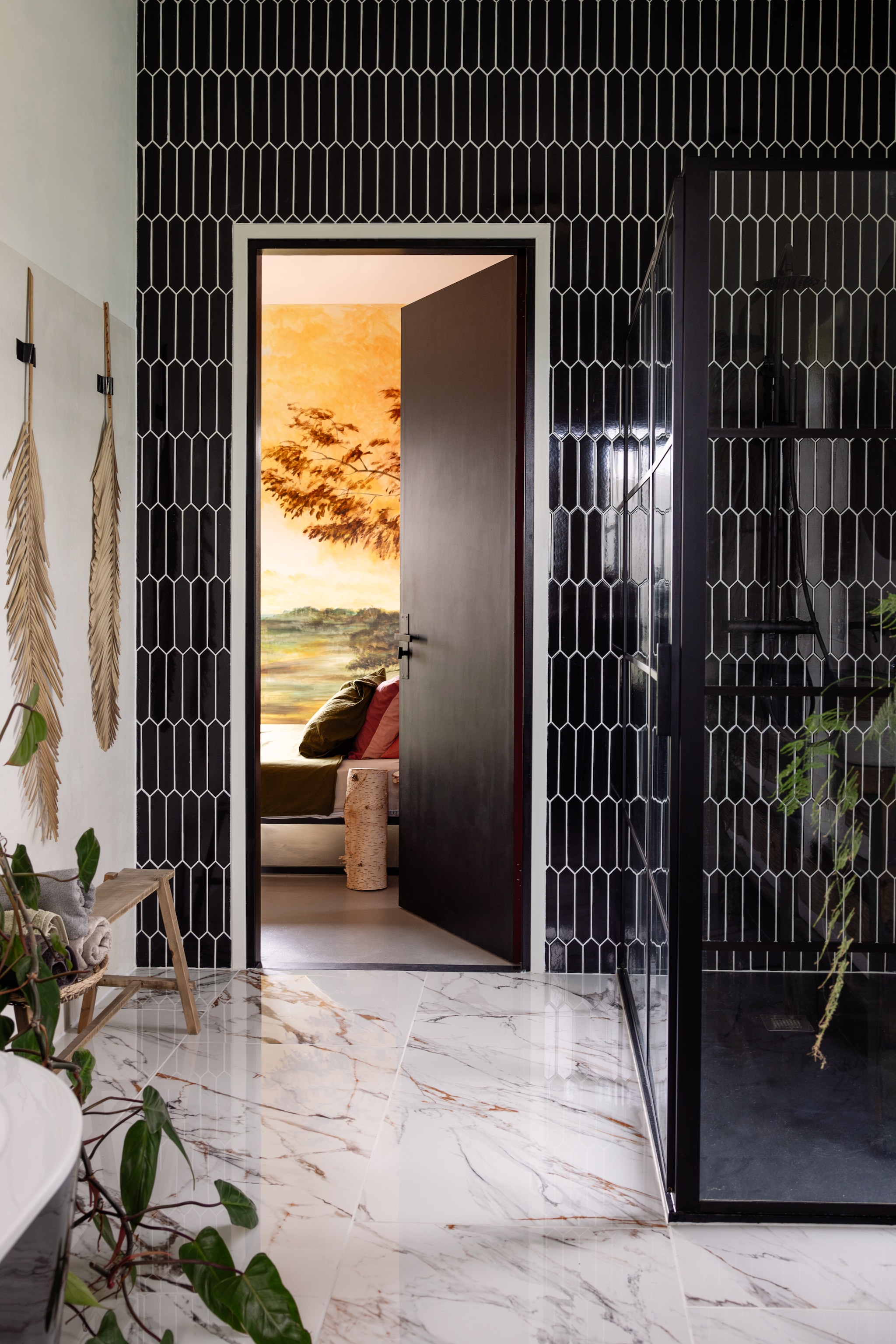 Vloertegels Verona Marmer en wandtegels Arrow Black van Tegels in Huis. Muurverf Beton ciré in wit, rotan bank Xenos, douchecabine Tegeldepot. Doorkijkje: muurschildering door Jessie gemaakt, bedframe VidaXL, beddengoed Dekbed Discounter, bijzettafel van berkenstam uit eigen tuin. vtwonen 02-2025 | styling ilona jongepier | fotografie dana van leeuwen | tekst Mirjam Rosema-Verhulst