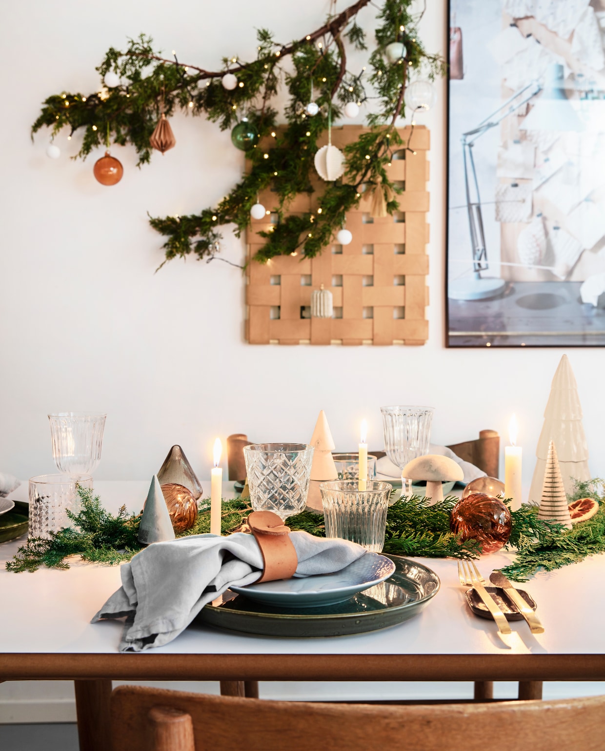 De kersttafel in het souterrain is eenvoudig maar stijlvol gedekt met sparrentakken,houten kerstboompjes en vintage kandelaars. Glazen van de vlooienmarkt, groeneborden van Bitz, kerstborden van Royal Copenhagen en de rommelmarkt. Tafel enbestek van Ilva, linnen servetten van Society, lamp van Anour. De servetringen zijnzelfgemaakt van leer. Ook het leren werk aan de muur is gemaakt door Rikke. Depijnboomtak is versierd met glazen ornamenten en zelfgemaakte papieren decoraties vtwonen 13-2019 | Styling Rikke Graff Juel | Fotografie Christina Kayser O.