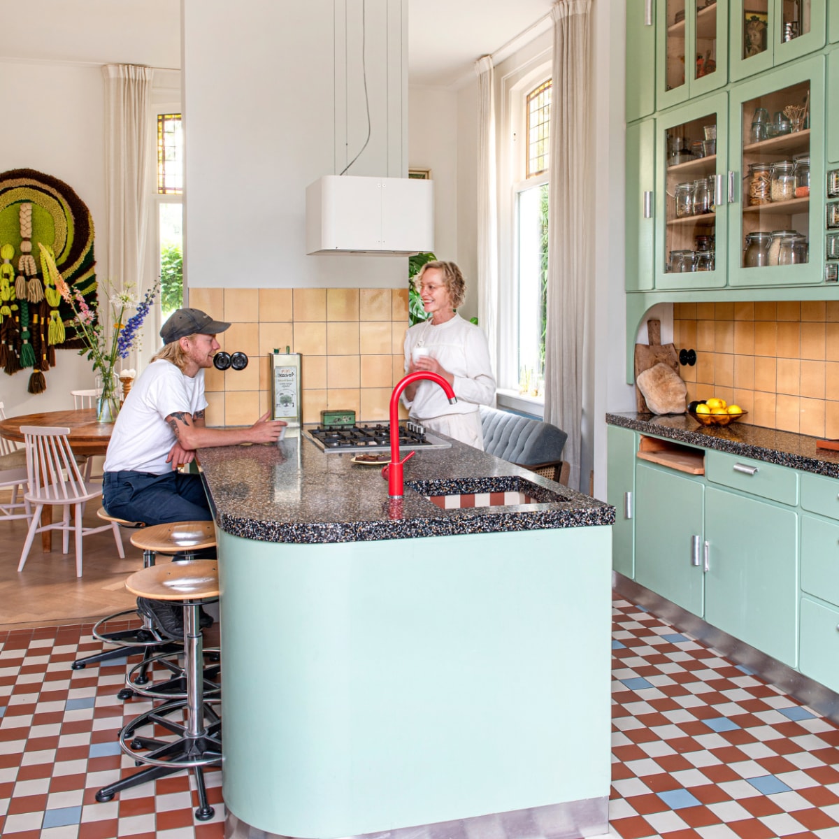 Keuken ontworpen door Studio Daan Hietbrink, verfkleur fronten Room Green 81 van Farrow &amp; Ball. Keukenblad Marcolina Terrazzo en Beton. Rode kraan sanitair.nl. Wandtegels en barkrukken Marktplaats. Vloertegels Winckelmans. Fruitschaal en broodplank uit Frankrijk. Overig onbekend. vtwonen 03-2023 | Styling en fotografie Caroline Coehorst i.s.m. Studio Daan Hietbrink