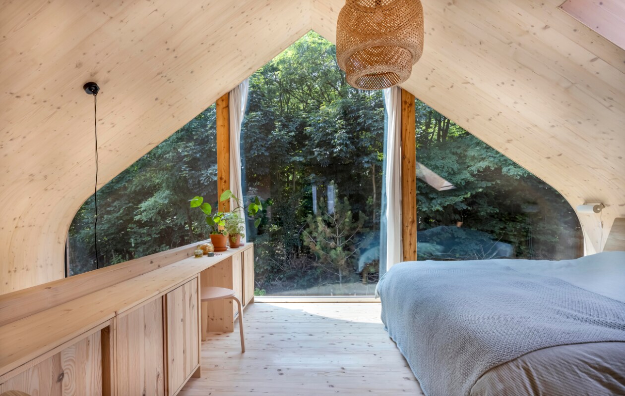 Ook de balustrade in de slaapkamer heeft kastruimte en een zitplek. Door de glasgevel komt het bos binnen. Hanglamp, groen beddengoed Ikea. Sprei Sissy-Boy, eiken bed Ethnicraft. vtwonen 01-2025 | fotografie Hennie van Belkom