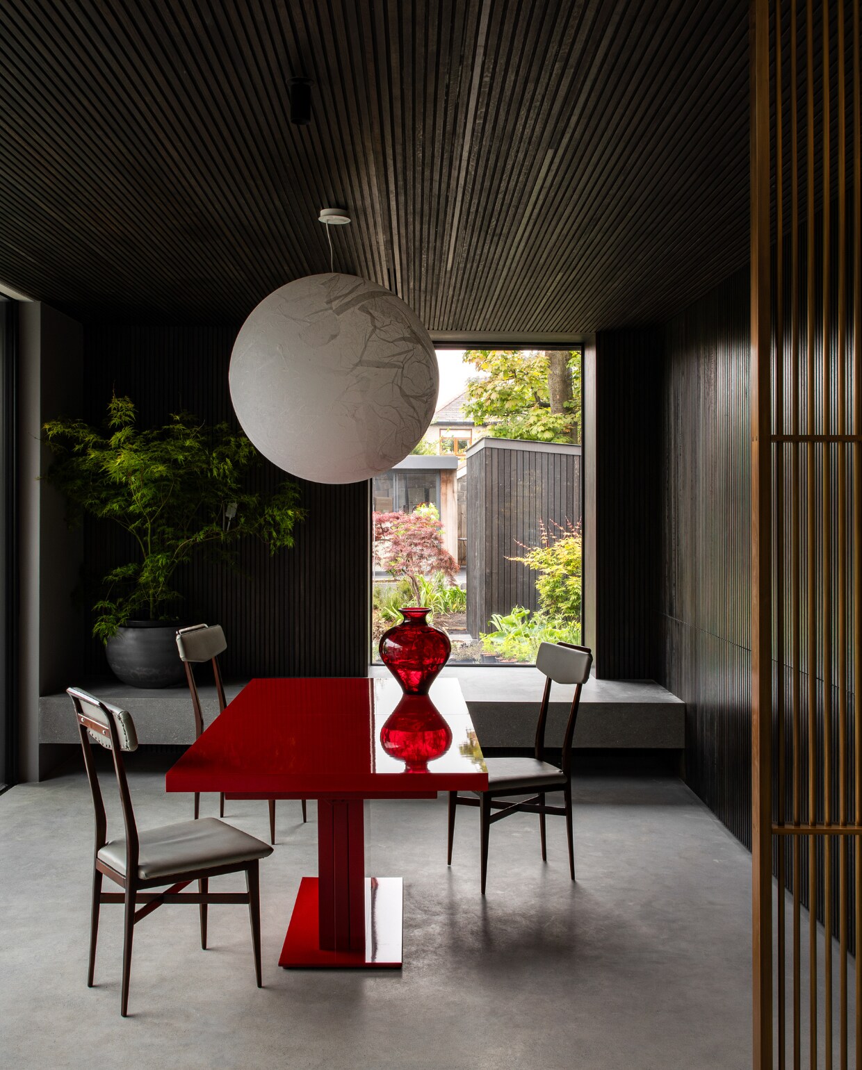 Vanuit de keuken kijk je naar de eetkamer. De donkere houten wandbekleding is gemaakt door het Dublinse bedrijf O’Gorman Joinery. De tafel had het gezin al en werd rood gespoten. Hanglamp van Davide Groppi, antieke stoelen Acquired. De bank voor het raam matcht met de betonnen vloer van Stone Seal. stijlvol wonen 01-2025 | productie Living Inside | fotografie Ruth Maria Murphy