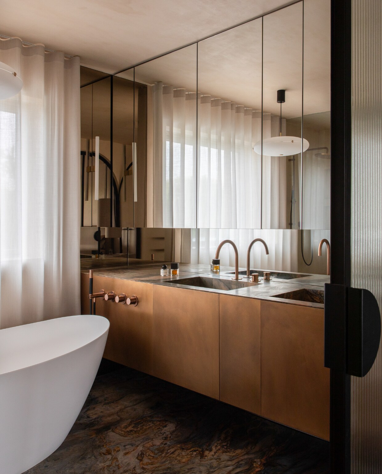 De badkamer bij de ouderslaapkamer kreeg een stenen vloer en werkblad van Miller Brothers. Koperen wastafelmeubel van Gessi, erboven een spiegelkast die wegvalt in de spiegelende spatwand. Dubbele wandlamp Nea van Kaia Lighting, hanglamp Luna Sospensione van Gio Ponti. stijlvol wonen 01-2025 | productie Living Inside | fotografie Ruth Maria Murphy