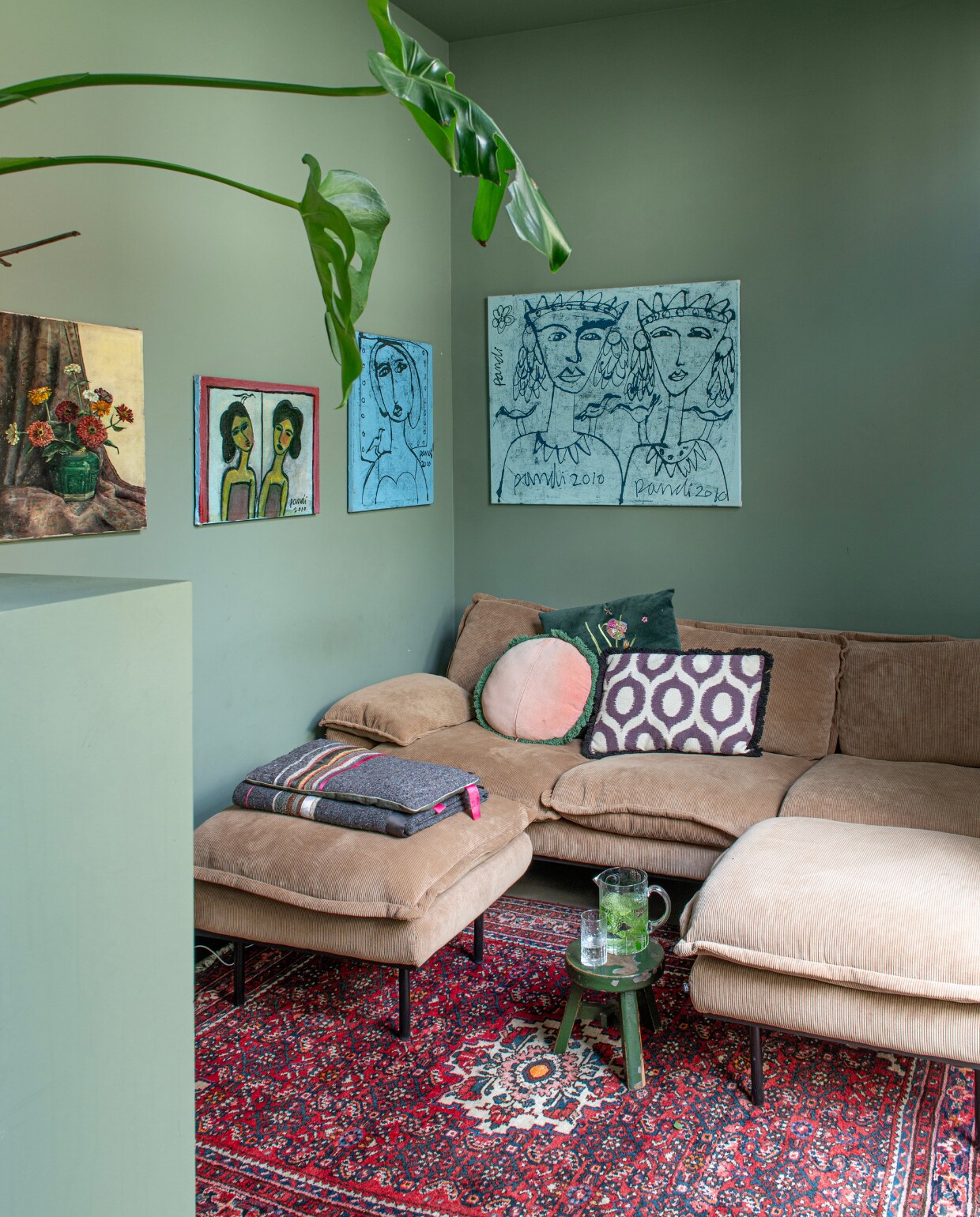 Bank HKliving. Kussens Sissy-Boy en Karwei. Plaid Ladak, portretten uit Indonesië, bloemschilderij kringloop,Perzisch tapijt van ouders geweest, kruk Sissy-Boy, karaf Iittala, muurverf Card Room Green van Farrow &amp; Ball. vtwonen 01-2025 | productie De Binnenkijkers | fotografie Caroline Coehorst