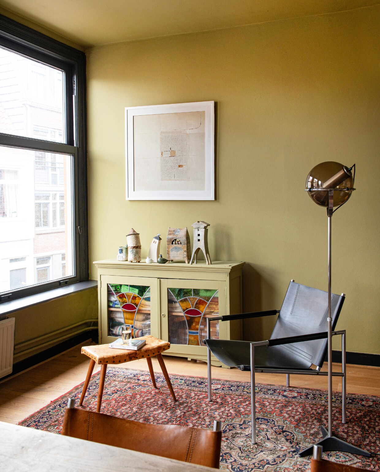 Muurverf Churlish Green van Farrow &amp; Ball. Kunstwerk uit ouderlijk huis Rogier, vintage fauteuil van Martin Visser, lamp Globe van Raak, kruk kringloop. Kast uit ouderlijk huis van Leonie, in kleur van de wand geschilderd. Objecten erop uit verzameling keramiek van Rogiers moeder. Perzich tapijt uit een  faillissements-verkoop. vtwonen 01-2025 | productie De Binnenkijkers | fotografie Caroline Coehorst