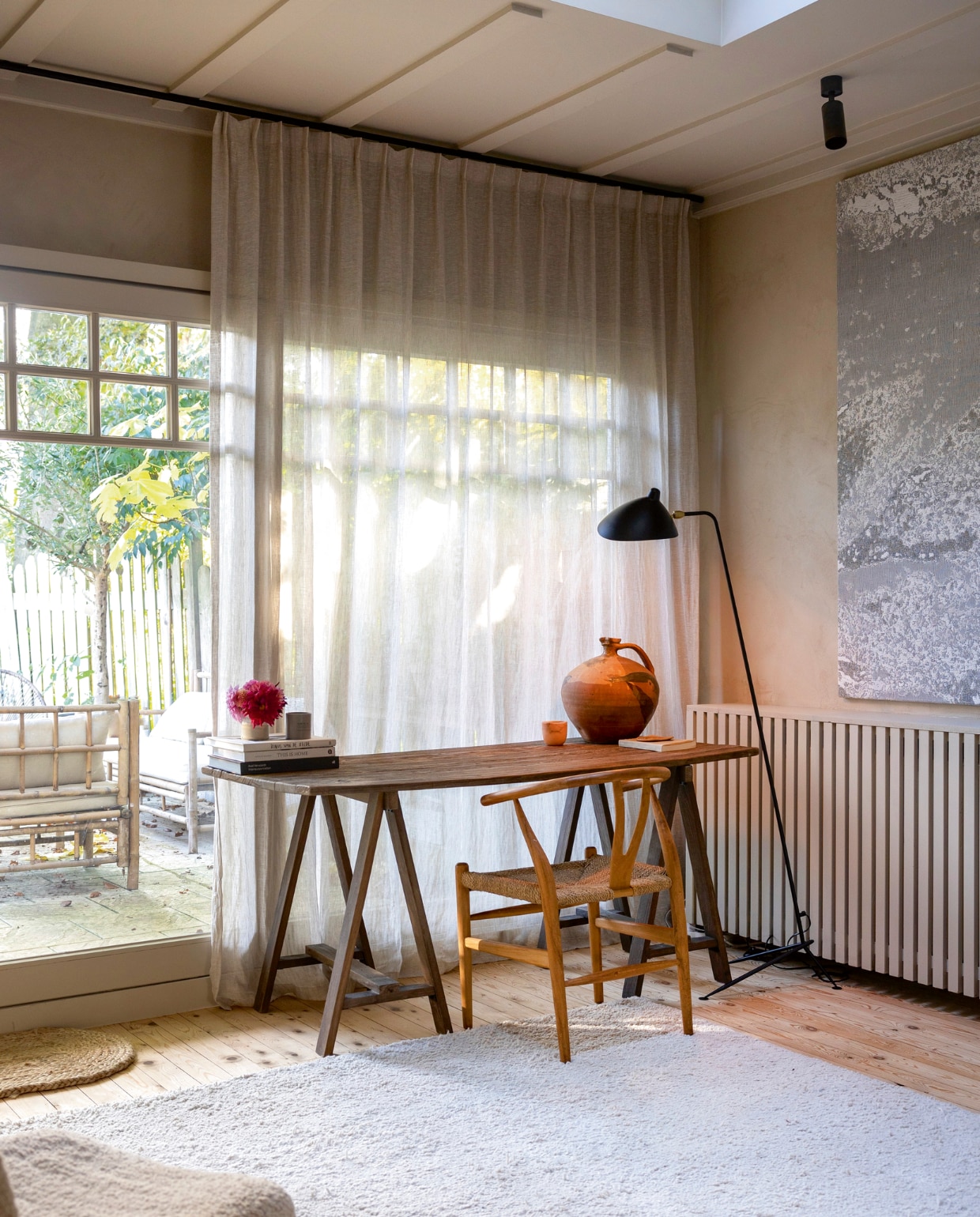 De werkplek met uitzicht op de tuin. Bureau van Zara Home, kunstwerk van Nathalie Van Der Massen, lamp van Serge Mouille. De halfhoge lattenwand is een op maat gemaakte  radiatorombouw. Stoel Wishbone van Carl Hansen. Kruik gevonden op een markt in Frankrijk. vtwonen landelijk 01-2025 | fotografie Sarah van Hove