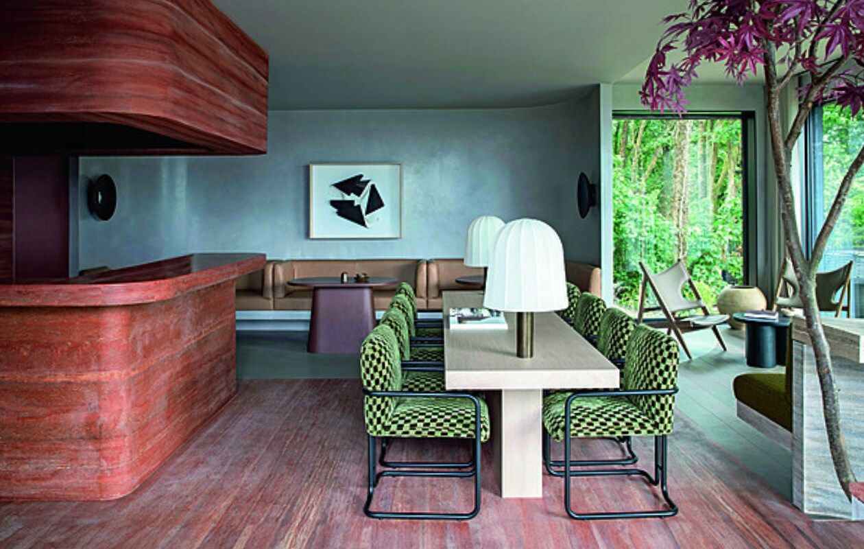 De Clubhouse voor de gasten van de Woodland Suites met centraal een eiken tafel. Op de achtergrond op maat gemaakte leren banken en roodbruine tafels. Kunstwerk van Manolo Ballesteros. Voor het raam staat de Knitting Lounge Chair van Audo Copenhagen. Stijlvol Wonen 01-2025 | productie Living Inside | fotografie Ruth Maria Murphy
