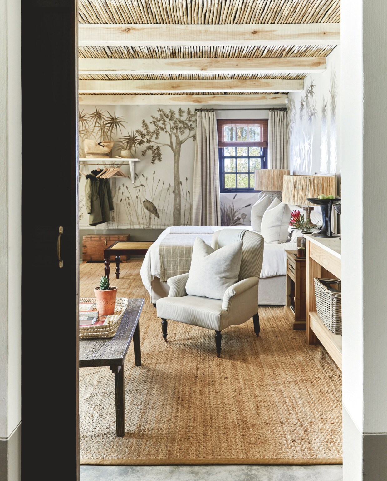 Deze kamer in het bijhuis heeft twee bedden, een kleine zithoek, een gietijzeren kachel voor koude winteravonden en meer van Sarah Pratts muurschilderingen. Soortgelijk sisaltapijt Wool and Wire, soortgelijke raffia lampenkappen Xenos. vtwonen landelijk 02-2025 | Styling Luanne Toms | Fotografie Elsa Young | Tekstbewerking Mirjam Rosema-Verhulst