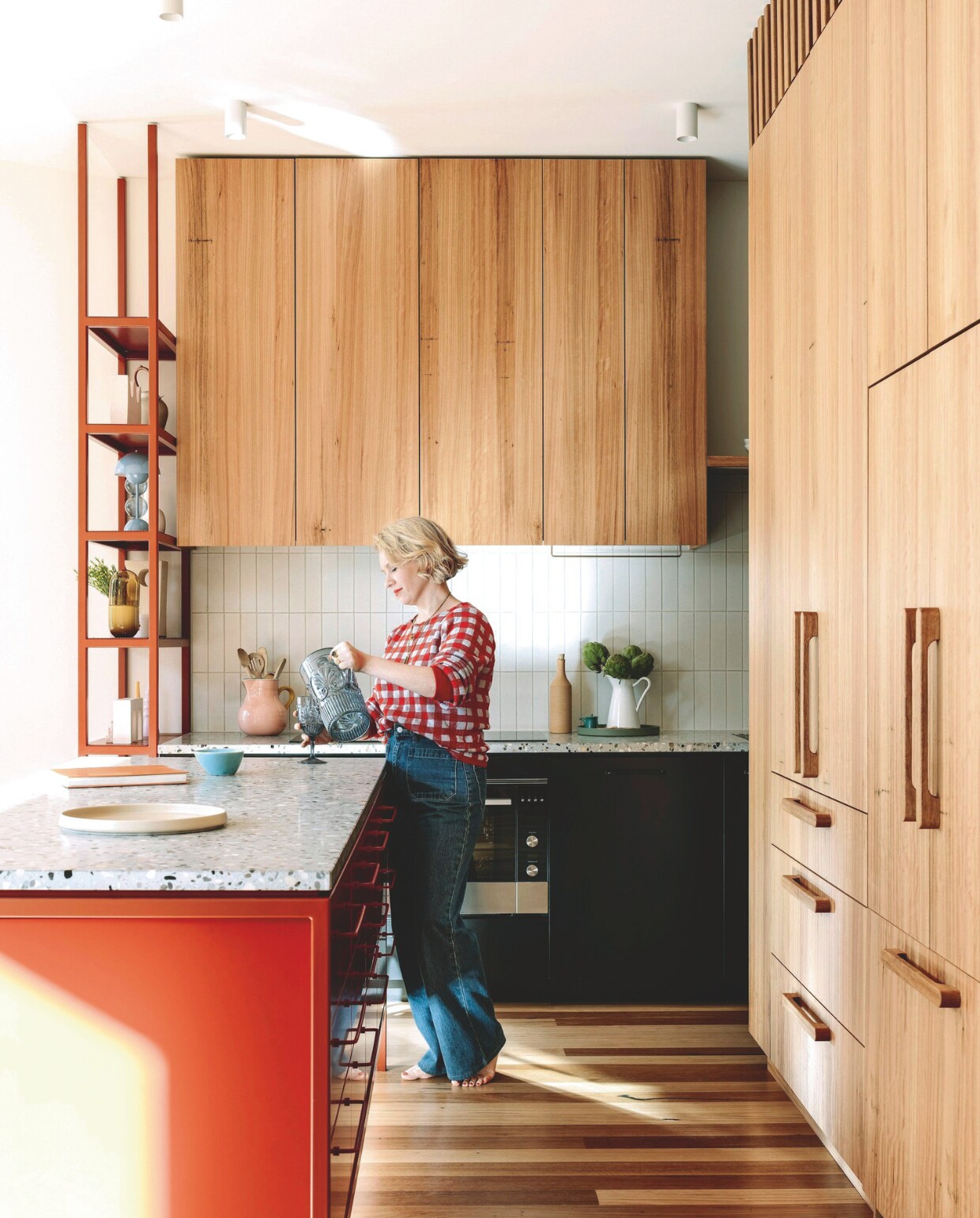 Keukenkasten, -eiland en hoge stellingkast, ontworpen door Imogen Pullar Architecture, gebouwd door J&amp;RC Builders met duurzame blackbutt-fineer. Handgemaakte houten handvaten Auburn Woodturning. Rood stalen frame en handgrepen Lounge van Kethy. Terrazzo keukenblad Moon Garden van Fibonacci. Tegels Hida van Academy Tiles. vtwonen 03-2025 | styling belle hemming | fotografie marnie Hawson | tekst caroline westdijk