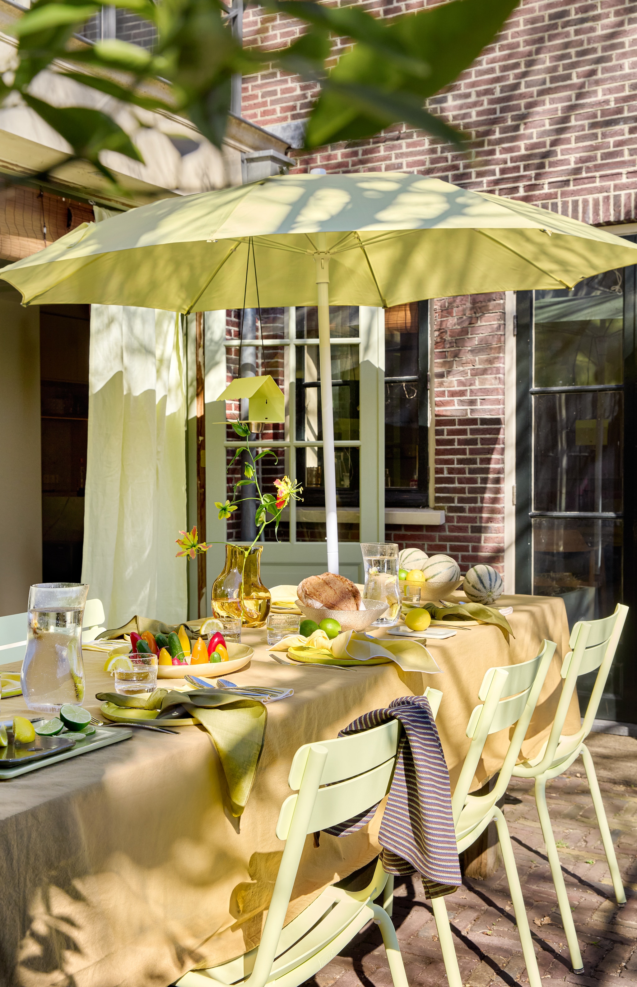 Aluminium tuinstoel Luxembourg van Fermob in Frosted Lemon € 269,-, parasol van Fermob in Frosted Lemon € 455,-, aluminium vogelvoederhuisje Picoti van Fermob in Frosted lemon € 65,- (extravert.nl). Katoenen tafelkleed Washed Mosterd van Kook € 39,99 (bol.com). Glazen vaas Gleam € 24,95 (hkliving.com). Linnen servetten van Kulu Club in olijfgroen en botergeel, set van 2 € 22,95, glazen waterkan Sui Bubbles Clear van Hübsch € 22,-, glazen Sui van Hübsch € 6,-, katoenen theedoek in bruin van Hübsch € 12,-, keramieken borden Amare in olijfgroen van Hübsch € 38,-, stalen bestek Mano van Hübsch € 100,-, glazen kom Fuyu van Hübsch € 8,50 (shop.vtwonen.nl). vtwonen 06-2025 | styling Kelly van Gendt | fotografie Peggy Janssen