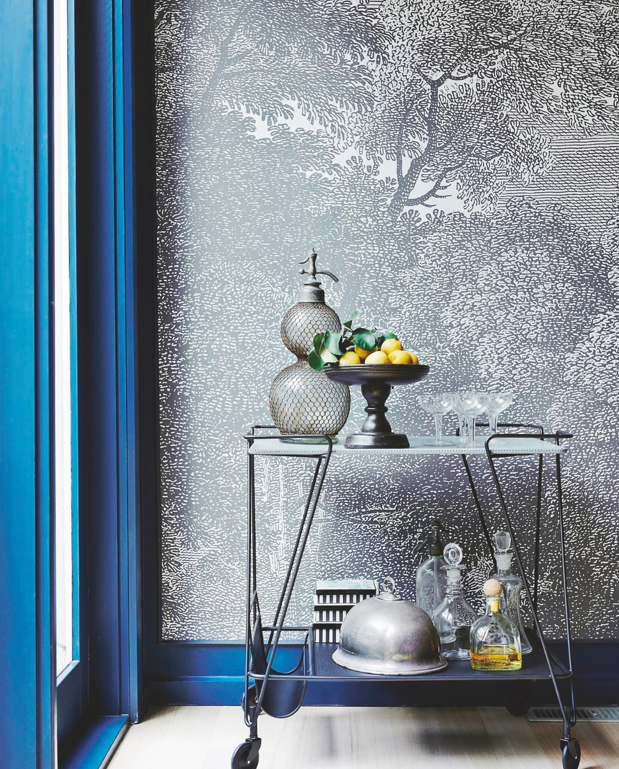 Muurkleur afgestemd op Hague Blue van Farrow &amp; Ball. Behang Arcadia van Anthropologie. Trolley van Gubi via Cult Design. Kristallen karaffen, frisdrankdispenser en cloche vintage. vtwonen 04-2025 | styling Fiona Richardson | fotografie Lisa Cohen