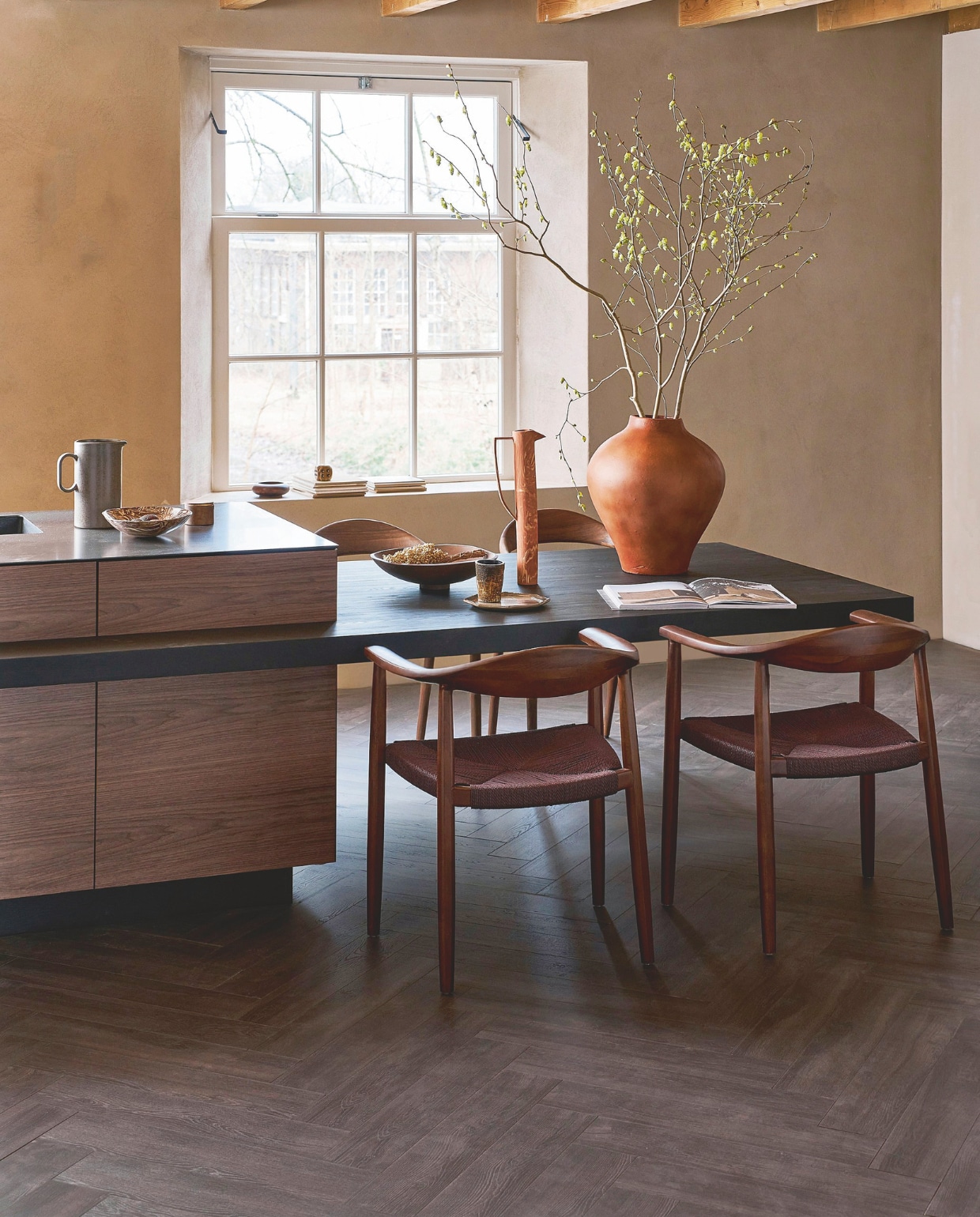 Laminaat visgraat in Collar Donkerbruin, lamel 65,5x13,3x0,8 cm, met groef, per m² € 37,99.M.m.v. shop.vtwonen.nl (eetkamerstoelen Embrace van vtwonen), mettesophie.com (hoge kan, aardewerken schaal), zarahome.com (bord), dofine.nl (wandafwerking), urbannatureculture.com (houten schaal, vaas). Overig privébezit. vtwonen 05-2025 | styling Cleo Scheulderman | fotografie Jeroen van der Spek