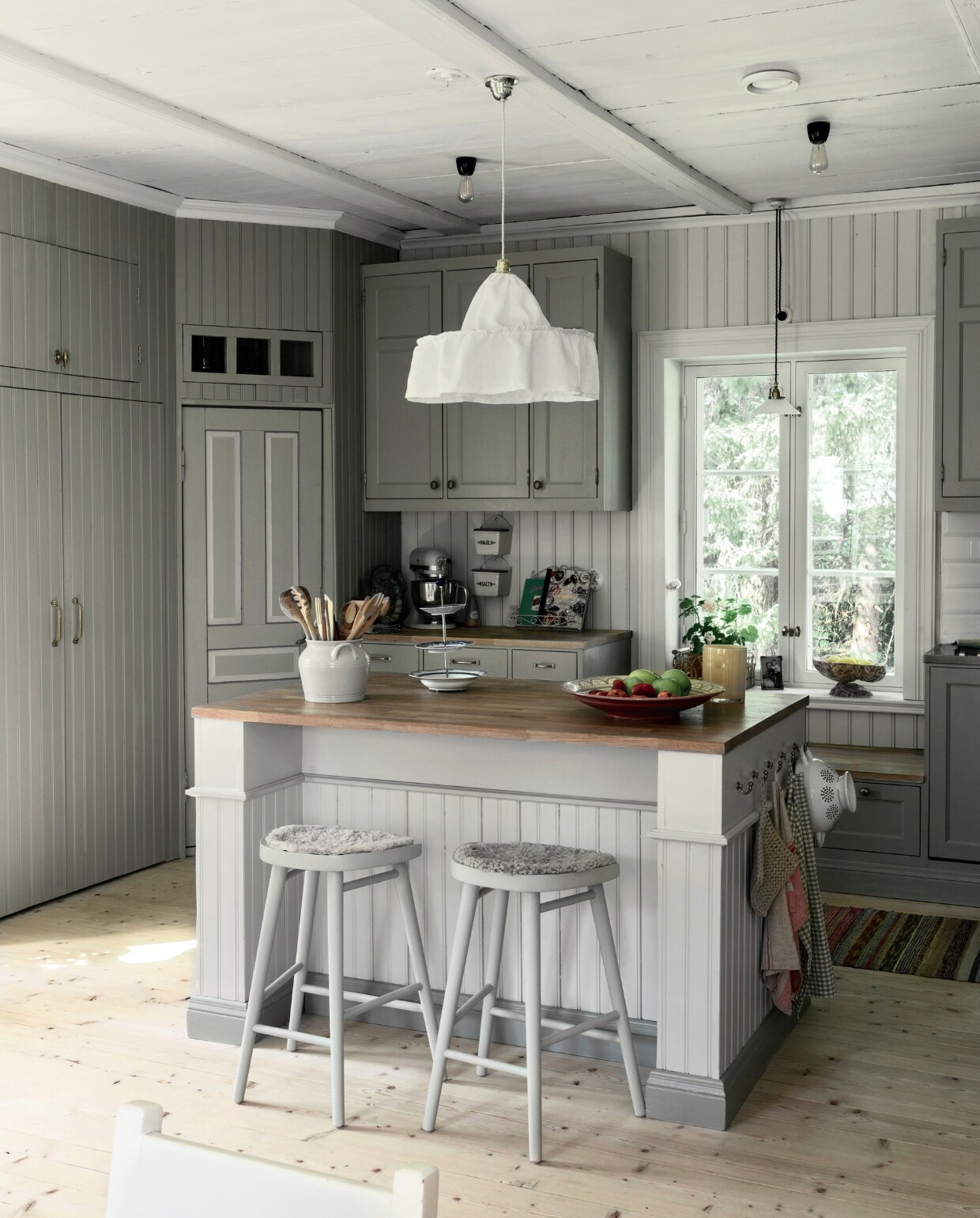 Het middelpunt van de keuken is het eiland. Mooi afgewerkt met panelen en sierlijsten, en volledig met de hand gemaakt.Het wordt verlicht door een lamp die Jenny zelf naaide, gebaseerd op een ontwerp uit het begin van de 20e eeuw (vaak de Carl Larsson- of Josef Frank-lamp genoemd). vtwonen Landelijk 06-2025 | styling, fotografie & tekst Heikki Rautio | productie Studio Rautio