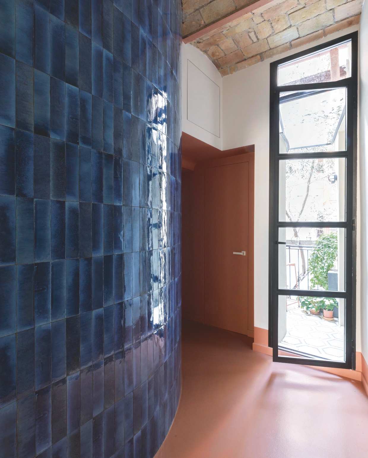 De gang naar het slaapgedeelte heeft een gebogen muur die geheel bedekt is met tegels, vergelijkbaar: bejmat-look Italy Blu via Tegelinfo. De terra gietvloer zorgt voor een modern contrast. Stijlvol Wonen 04-2025 | styling Francesca Sironi | fotografie Monica Spezia | tekstbewerking Tanne Donjarcour Gielen | prouctie Living Inside | tekst Francesca Sironi