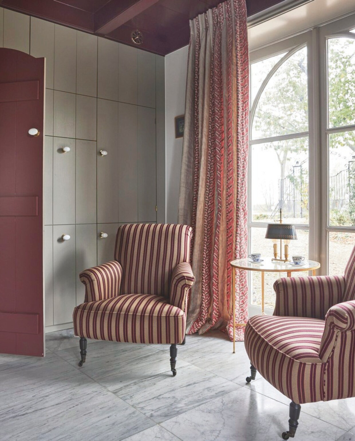 Fauteuil Bonifacio in stof Rayure Gladstone Nobilis, via Wilhelmine van Aerssen Agenturen. Bijzettafel Vaughan Designs. Antieke Bouillot tafellamp. Kastenwand op maat gemaakt door Hans Tinga. Kleur houtwerk Persiaans rood van Vettewinkel. vtwonen landelijk 01-2025 | styling Marianne Luning | fotografie Alexander van Berg