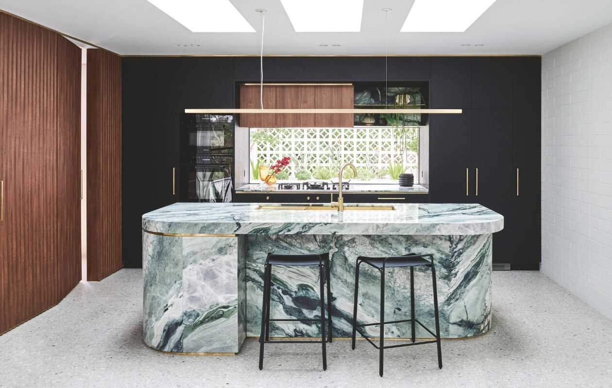 Hanglamp Giffin Design. Het Calacatta Verde-marmer voor het eiland kreeg ronde hoeken in de looproute, en messing details die de belijning versterken. Kasten maatwerk.  Deurbeslag Designer Doorware. Wandbekleding Screenwood. Krukken StyleCraft. Op de vloer ligt terrazzo. Dakramen Velux. Stijlvol wonen 05-2025 | fotografie Tom Blachford | tekstbewerking Evelien Roels