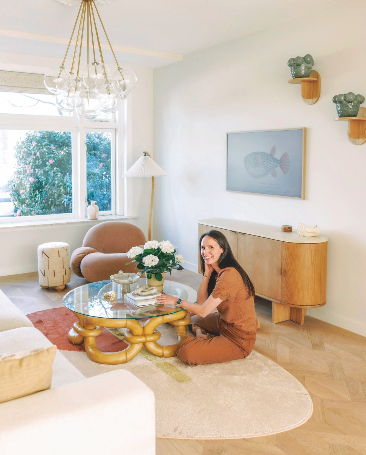 Salontafel Dusty Deco Bamboo Bracelet Coffee Table via Studio Uma. Dressoir, schaal op pootje, houten muurplanken en vaas in vensterbank Westwing. Witte schaal cadeau. Onderzetters van Michelles grootouders uit Indonesië. Televisie Samsung The Frame. Keramieken vazen Mica Decorations. Fauteuil Gubi Pacha in stof Dedar. Staande lamp Gubi. Hanglamp Eichholtz. Poef Amstelièr. Eiken parketvloer Vloerenhuis Amsterdam. Kleed Zuiver. Ornamenten plafond Het Schippertje. Vouwgordijnen Studio Diezijn.  vtwonen 7-2025 | fotografie Emma Peijnenburg