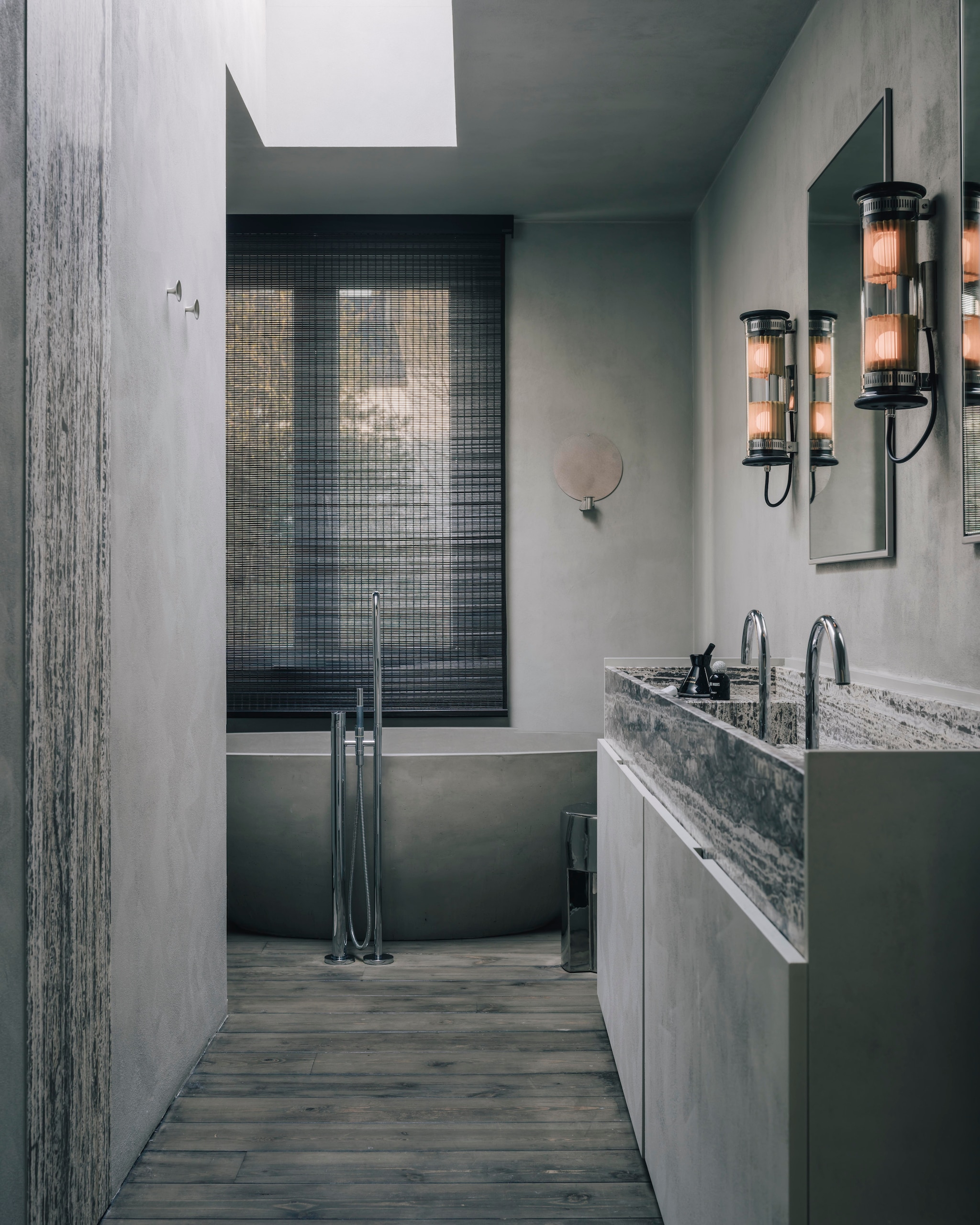 In de serene badkamer voeren grijstinten de boventoon. Wandlampen In The Tube. Naast het cementen bad een vrijstaande kraan van Vola. Erboven een shutter van zwart bamboe en een rond ornament voor een kaars van Frama. Vooraan de op maat gemaakte wastafel van travertin met wastafelkranen van Hotbath. Stijlvol wonen 9-2025 | Katoo Peeters