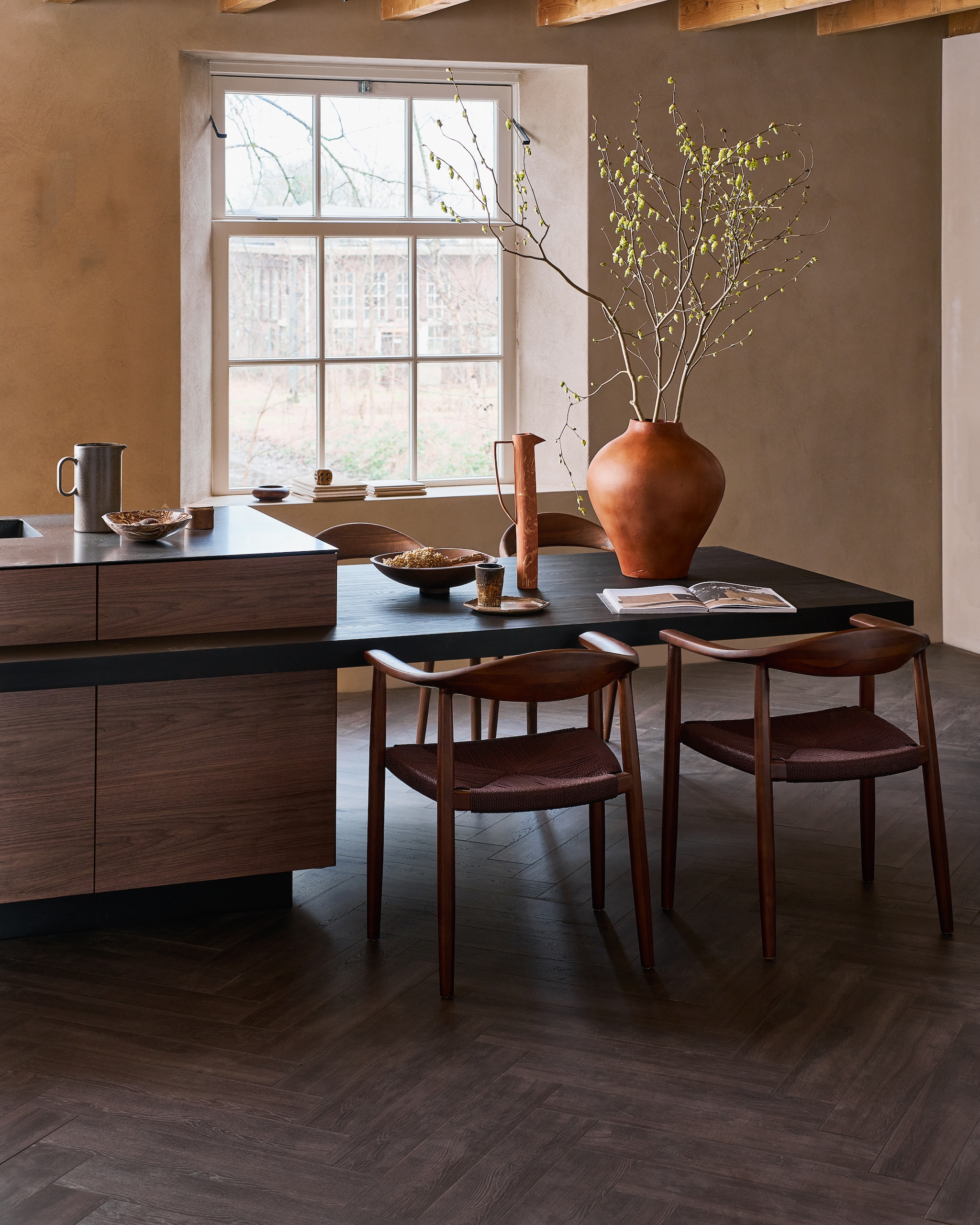 Laminaat visgraat in Collar Donkerbruin, lamel 65,5x13,3x0,8 cm, met groef, per m² € 37,99. M.m.v. shop.vtwonen.nl (eetkamerstoelen Embrace van vtwonen), mettesophie.com (hoge kan, aardewerken schaal), zarahome.com (bord), dofine.nl (wandafwerking), urbannatureculture.com (houten schaal, vaas). Overig privébezit. vtwonen 05-2025 | styling Cleo Scheulderman | fotografie Jeroen van der Spek