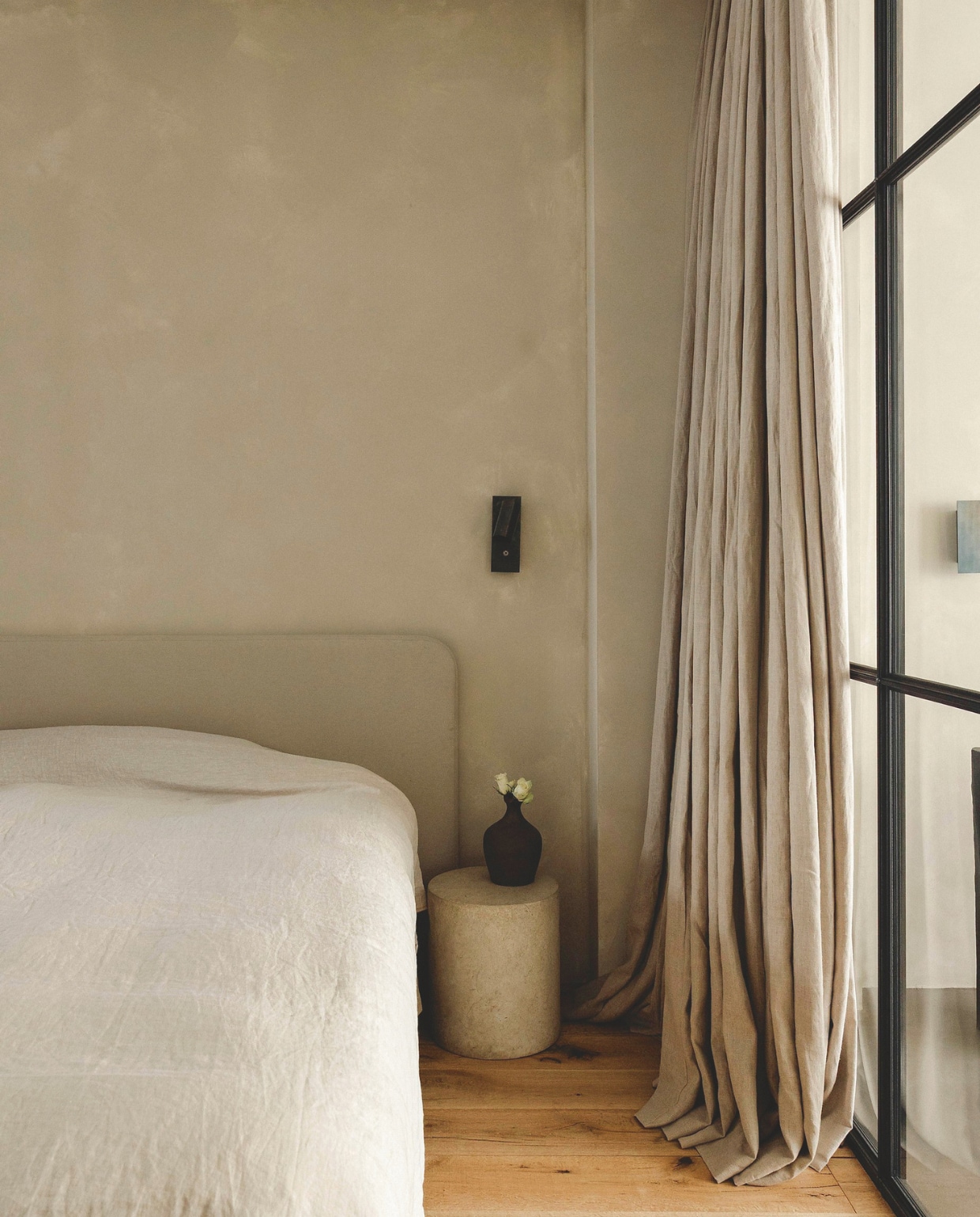Linnen hoofdbord op maat gemaakt De Slaapkamer. Linnen sprei Zara Home. Nachtkastje Pomax. Vaas antiek. Wandlamp Serax. Gordijnen op maat gemaakt Kwantum. Houten vloer Wood and Stones. Op de muur betonverf in Silk van Beton Cire Webshop. vtwonen landelijk 8-2025 | fotografie Stie De Neve