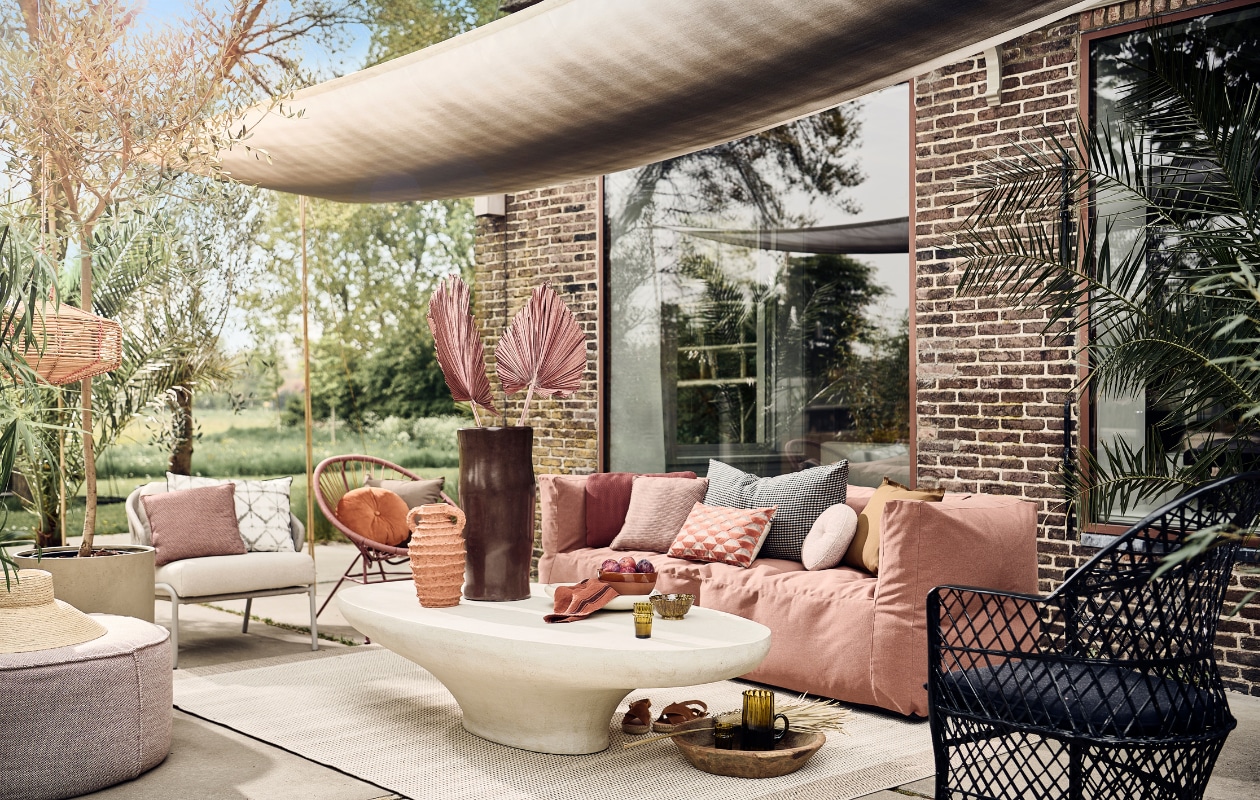 Schaduwdoek gemaakt van buitenstof Padua van Designers Guild, per meter € 73,-, wit rechthoekig buitenkussen met ikat print € 70,- (wva.nl). Buitenpoef vanaf € 525,-, zachtgrijze fauteuil met witte zitting € 2620,-, terra vierkante tuinkussens, per stuk € 151,-, terra langwerpig tuinkussen € 151,-, zachtroze rond tuinkussen vanaf € 410,- (dacks.nl). Hoed € 39,99 (zarahome.com). Rotan windlicht € 39,99, terracotta schaal, 20 cm (Ø) € 29,99, suède sandalen € 79,99 (sissy-boy.com). Betonnen bloempot vanaf € 150,- (nordal.eu). Buitenkleed van Lafuma, 200×290 cm € 269,90, oranje rond buitenkussen van Hartman € 24,95, buitenkussen met oranje retro dessin van Hartman € 29,95 (vtwonen.nl). Bruine tadelakt vaas € 115,- terracotta vaas, 35 cm (h) € 85,-, witte saladeschaal, terracotta met tadelakt finish, 38 cm (Ø) € 47,50, mosterdgele kom van klei, 13 cm (Ø) € 12,-, glas, per stuk € 6,50, karaf € 39,-, polyester zwarte fauteuil € 429,- (householdhardware.nl). Microcement salontafel vanaf € 1499,- (lanena-home.com). Driezits buitenbank van Subrella stof, 223x80x75 cm (lxbxh) € 1450,- (bryck.nl). Geruit zwart beige kussen € 90,-, honingbruin buitenkussen met rafelrand € 25,-, beige buitenkussen met rafelrand € 25,- terra linnen theedoek € 11,- (paviljoennoord.com). Roze loungestoel Samantha van Kave Home, polyester en staal, 84x72x75 cm (hxbxd) € 126,- (shop.vtwonen.nl).